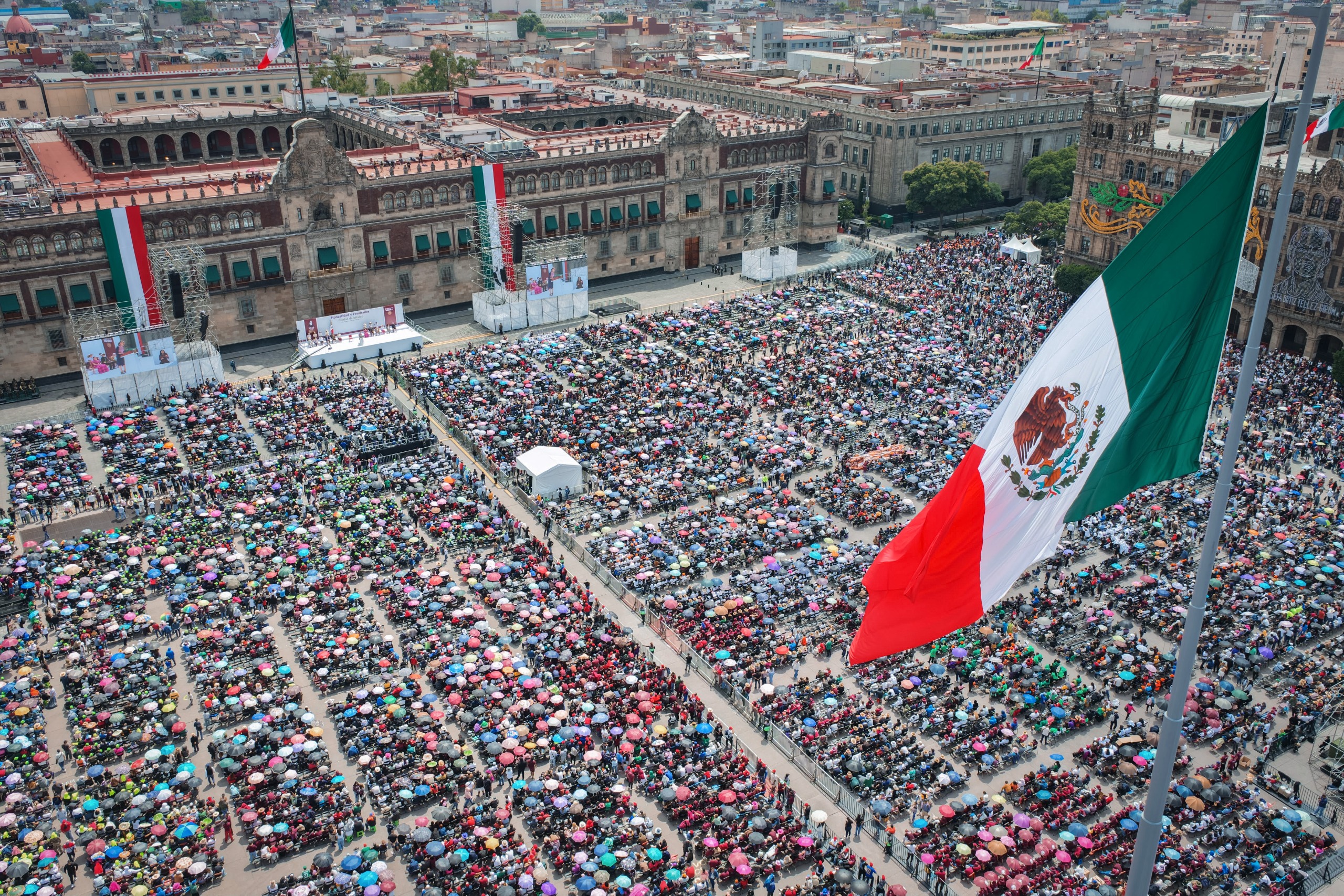 Vista panorámica del Zócalo con asistentes al acto y la bandera de México en primer plano.