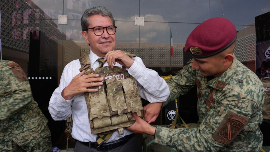 Ricardo Monreal ajusta un chaleco táctico con ayuda de un militar durante una exhibición.