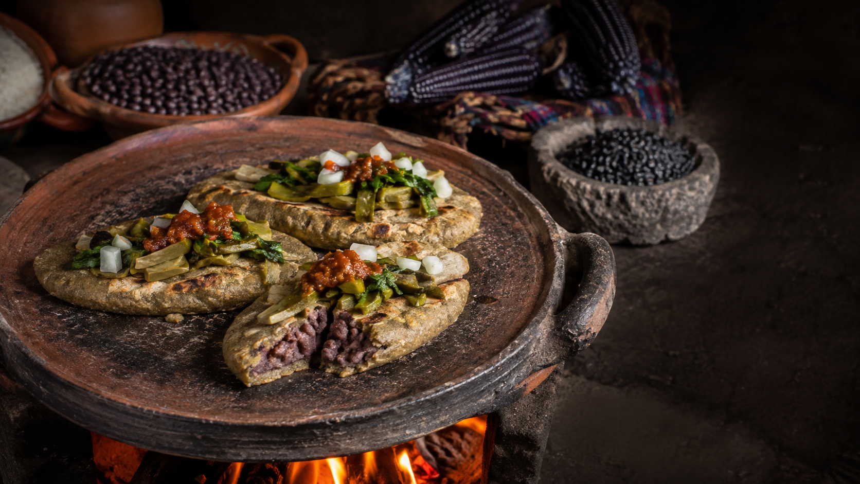 Tlacoyos de maíz azul rellenos de frijol y cubiertos con nopales y quelites, cocidos en un comal de barro tradicional.