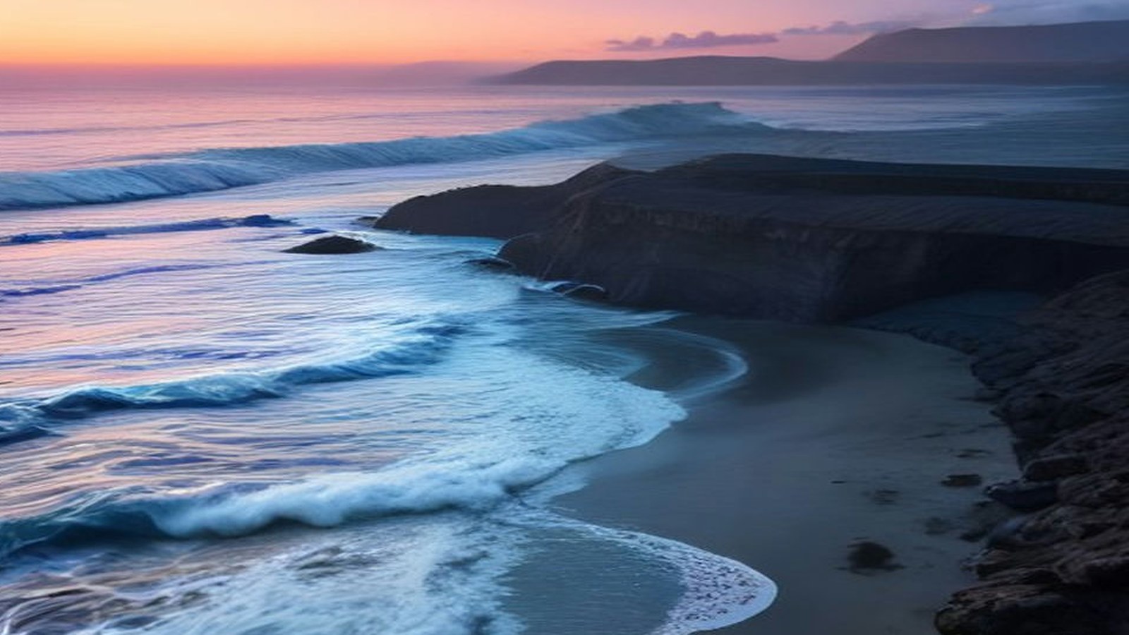 Olas del océano que rompen en una playa de arena oscura junto a acantilados rocosos al atardecer